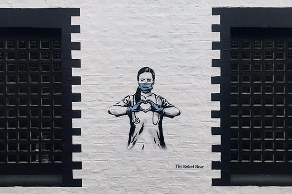 Street art by The Rebel Bear depicting a nurse in black and white with a blue face mask and gloves, with her hands making the shape of a heart.